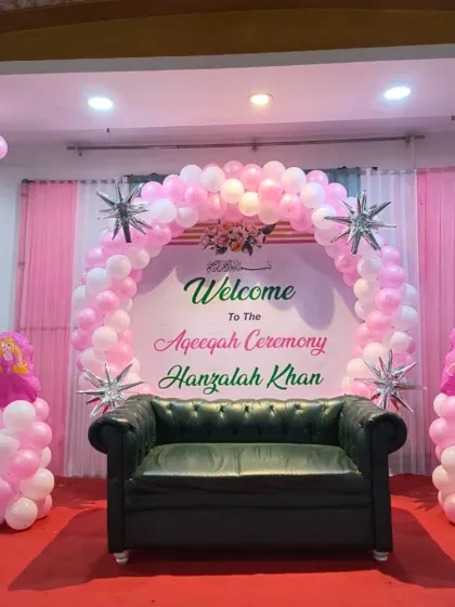 A beautiful stage for the "Aqeeqah Ceremony of Hanzalah Khan." The setup features a white and pink balloon arch over a sofa, with a personalized welcome sign on the backdrop.
