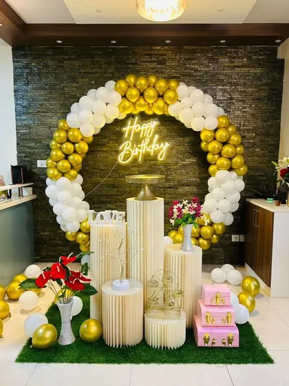 A luxurious birthday setup with a gold and white circular balloon arch against a dark, textured wall. The scene is set on artificial grass and includes multiple pedestals with flowers and props for a complete look.