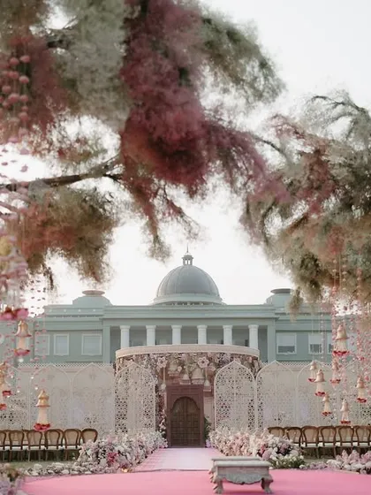 A grand mandap at Raffles Udaipur, with trees adorned in pink and white florals. The design creates a fairytale-like atmosphere, framing the palace in the background.