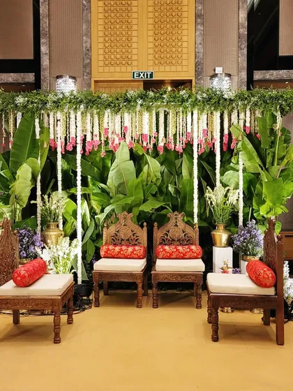 An elegant indoor setup for an engagement or pre-wedding ceremony, using traditional carved chairs, banana leaf backdrops, and hanging floral strings.