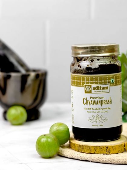 A jar of premium Chyawanprash is styled on a wooden slice with fresh amla and a mortar and pestle. The clean, bright setup emphasizes the product's Ayurvedic roots and natural purity.