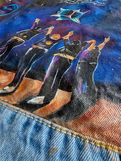 A close-up on the lower part of the jacket, highlighting the painted figures of the band members with their arms raised. You can see the texture of the acrylic paint on the denim fabric.