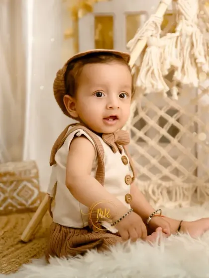 A baby boy in a stylish, neutral-toned outfit and cap, sitting in a boho-themed setup with a macrame teepee and pampas grass.