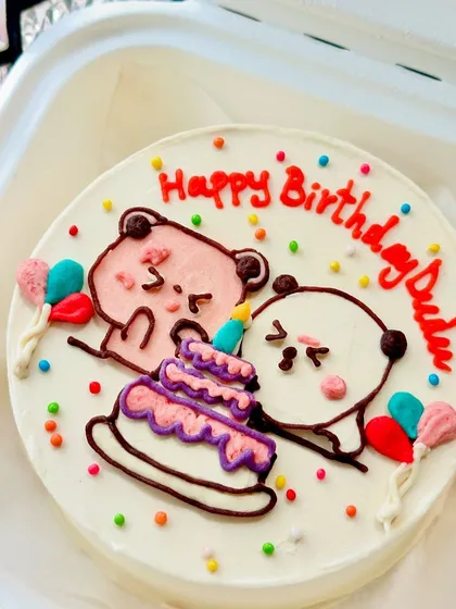 An adorable bento cake with two cute characters celebrating a birthday. The colorful sprinkles and tiny balloons add to the festive feel.