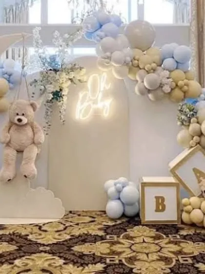 A dreamy teddy bear baby shower setup featuring a large moon prop and "BABY" blocks. The soft blue, beige, and white balloons, combined with the "Oh Baby" neon sign, create a magical and serene atmosphere.
