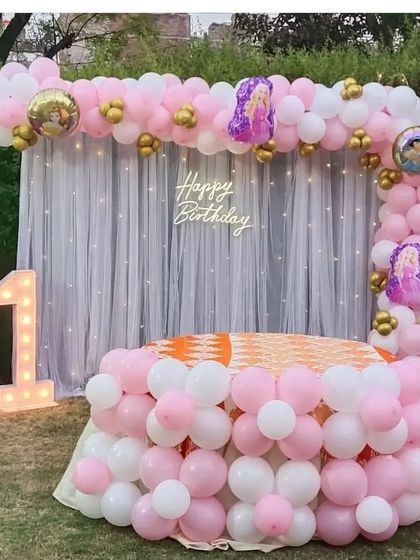 An outdoor first birthday party with a pink and white balloon arch, a "1" marquee number, and a sheer curtain backdrop with fairy lights.