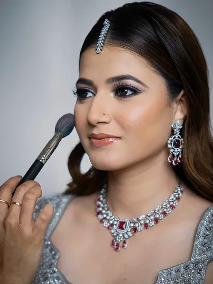 A behind-the-scenes moment showing a makeup artist applying blush. The model is wearing a stunning ruby and zircon necklace, giving a sneak peek of the glamorous final look.