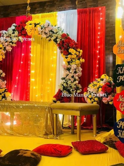 A colorful Mehendi setup with an arch of red, yellow, and white flowers, sequin drapes, and fun props.