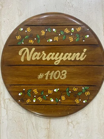 A custom hand-painted name board for a home entrance. This circular wooden plaque features the family name "Narayani" surrounded by a delicate floral vine.
