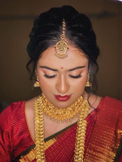 A classic South Indian bridal portrait. The makeup is centered around a bold red lip and defined, kohl-rimmed eyes, creating a timeless look that perfectly matches the richness of her red silk saree and gold jewellery.