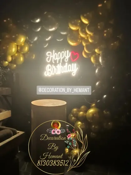 A moody and elegant birthday setup with a "Happy Birthday" neon sign. The dark setting is lit by the sign and a partial arch of gold and black balloons, creating a sophisticated ambiance.
