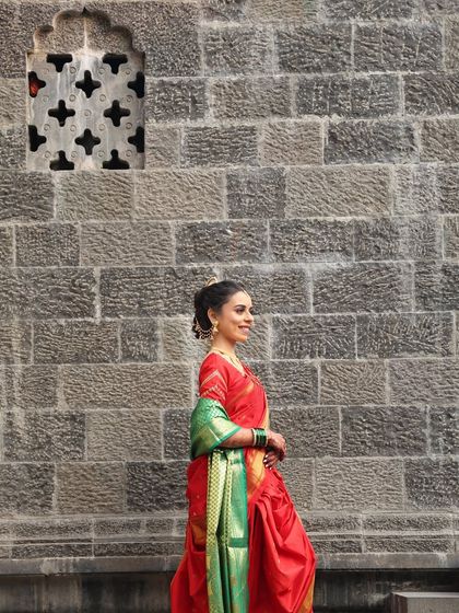 A stunning wide shot of the bride against a traditional stone wall. The vibrant red and green of her saree stand out, and her makeup ensures she looks perfect even from a distance.