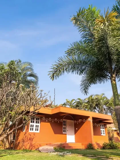 The exterior of a cottage at Vijayshree Resort, Hampi, surrounded by lush trees and greenery. The architecture reflects a simple, earthy style that blends with the natural landscape.