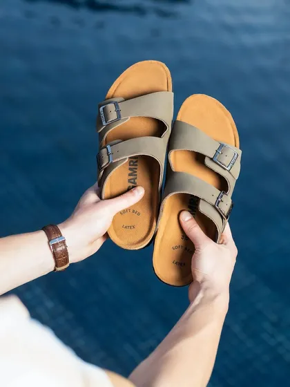 A point-of-view shot of someone holding a pair of sandals over the water. The bright, sunny lighting and clear blue background create a perfect summer vacation vibe.