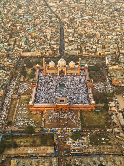 An aerial photograph capturing the golden hour light over Jama Masjid during Eid. The warm light beautifully illuminates the red sandstone architecture and the vast crowd gathered for prayer.