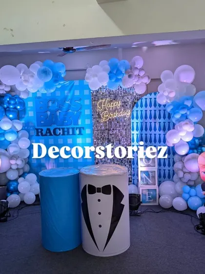A closer look at the Boss Baby themed decor, highlighting the custom backdrop with the child's name, the tuxedo-themed pedestal, and the detailed balloon work in shades of blue and white.