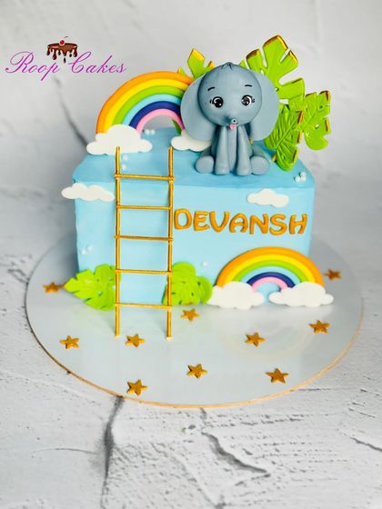 A cute half-birthday cake for Devansh. This design features a baby elephant on a ladder reaching for a rainbow, surrounded by stars and clouds.