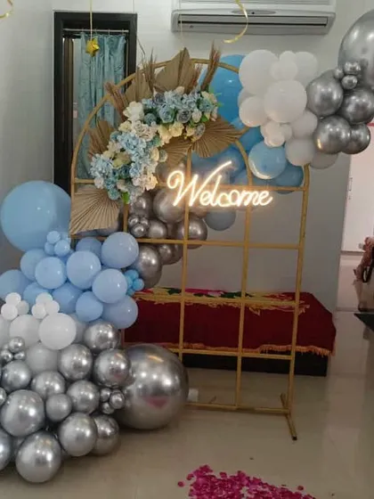A stylish welcome decor piece. The gold grid frame is decorated with a blue, white, and silver balloon arrangement and a "Welcome" neon sign.