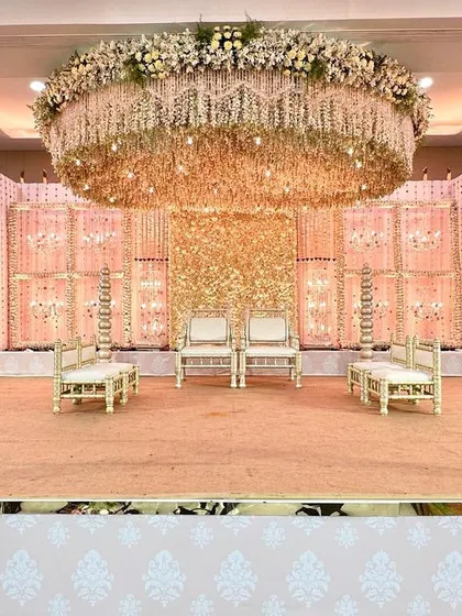 A full view of a Vidhi Mandap designed in soft pastel tones. The stage features a ceiling completely covered in hanging flowers and a backdrop of shimmering gold sequins, creating a space that feels both traditional and dreamlike.