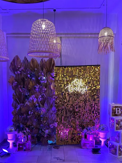 A glamorous baby shower setup in a restaurant, featuring a gold sequin wall, a floral wall, and an "Oh Baby" neon sign, proving any space can be transformed.