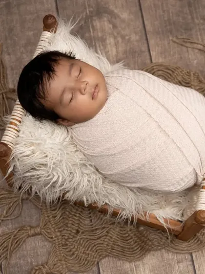 A sleeping newborn rests on a miniature charpoy, a traditional woven bed. The rustic wooden floor and simple swaddle create a timeless, culturally inspired portrait.