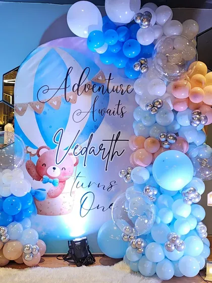 A close-up of the 'Adventure Awaits' first birthday backdrop. The design features a cute bear in a hot air balloon, surrounded by a beautiful pastel balloon arch.