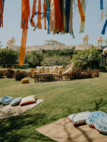 A relaxed, picnic-style seating area on the lawn. Mats and printed cushions invite guests to unwind under a tree adorned with flowing ribbons, with the grand Udaivilas palace in the background.