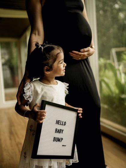 A quiet, observant moment. The older sister holds a "Hello Baby Bump" sign, looking thoughtfully at her mom's belly, anticipating the arrival of her new sibling.