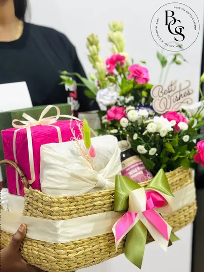 A large, impressive gift basket being held, ready for delivery. It's overflowing with fresh tuberoses and pink flowers, beautifully wrapped gifts, and a "Best Wishes" sign, perfect for any major celebration.