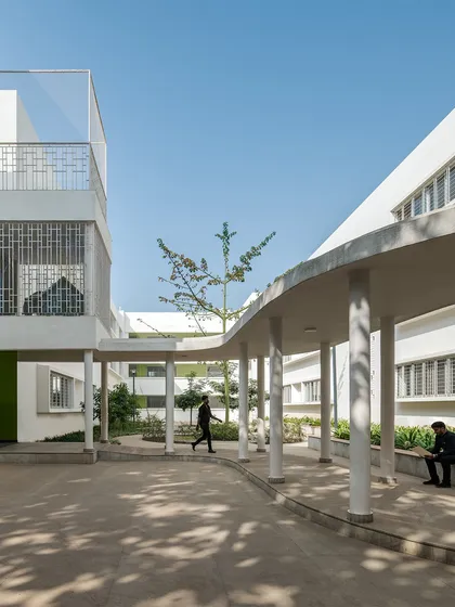 A curved, covered walkway at Vidyashilp Academy provides a shaded path between buildings. The organic form of the walkway contrasts with the rectilinear school blocks, creating a gentle and intuitive circulation route through the landscaped campus.