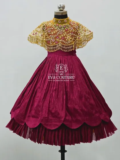 A unique wine-colored gown with a scalloped, layered skirt. It is paired with a stunning detachable cape featuring intricate gold maggam work.