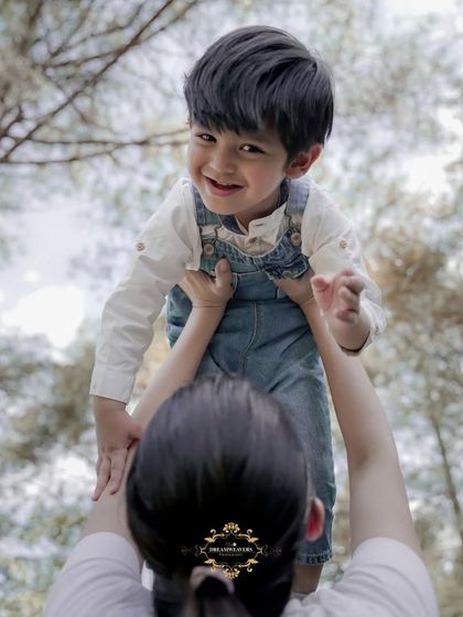 A mother lifts her son high in the air against a backdrop of tall trees. This low-angle shot captures a feeling of pure happiness and freedom.