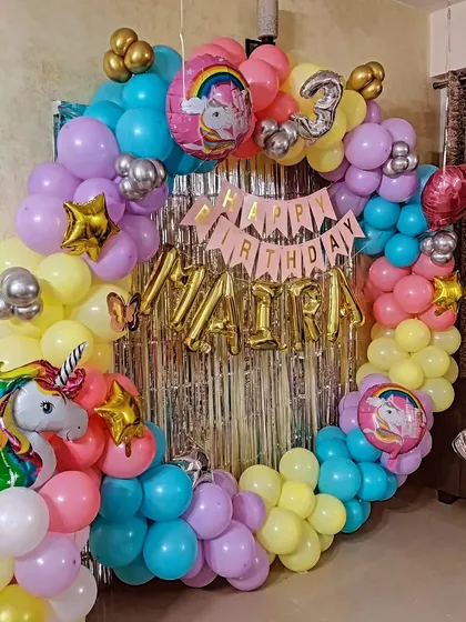 A pastel-colored unicorn ring decoration for a third birthday. The setup includes a silver fringe curtain, the child's name in foil balloons, and unicorn-themed foil balloons.