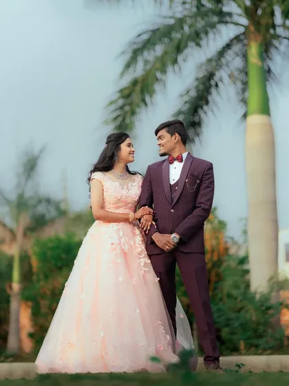 A classic garden engagement shoot. The bride-to-be looks lovely in a peach-pink ball gown with floral details, standing with her handsome partner.