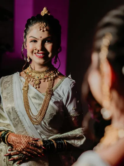 A classic bridal portrait showing the bride looking at her reflection. The lighting highlights her intricate gold jewelry and her happy, confident smile.