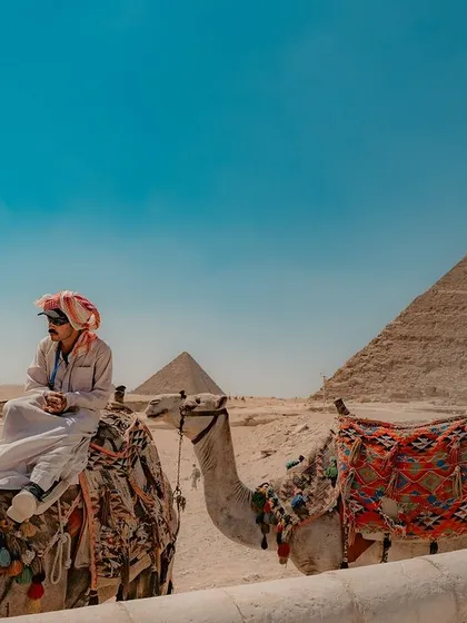 A man in traditional Arab attire sits atop a camel, with a pyramid in the background. This portrait captures a moment of quiet dignity against an iconic backdrop.