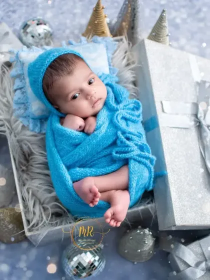 A winter-themed newborn photo where the baby is wrapped in blue and placed in a gift box surrounded by silver Christmas ornaments and props.