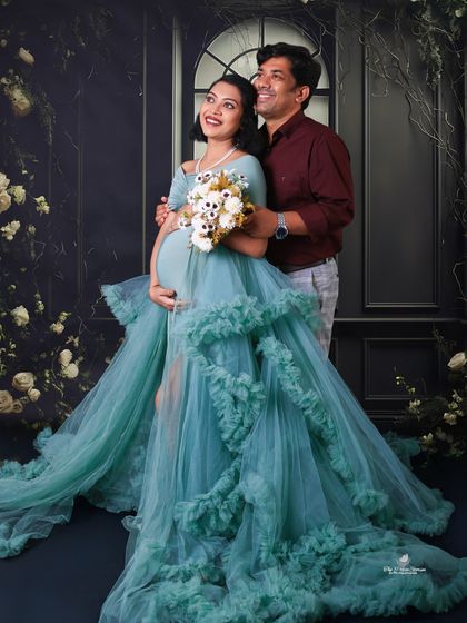 A dreamy and elegant solo portrait. The mom-to-be holds a bouquet, her teal gown flowing beautifully against a dark, romantic floral backdrop.