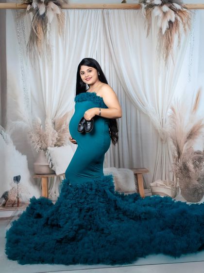 A smiling mother-to-be in a teal gown holds up a tiny pair of baby shoes in front of her bump.