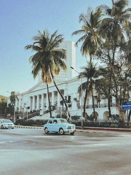 A light blue vintage Fiat drives past the Asiatic Society Library, with palm trees framing the shot. It's a perfect, sunny Bombay scene.