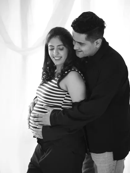 A candid black and white photo of a couple sharing a happy moment. The partner embraces the mother-to-be, both looking down at the baby bump.