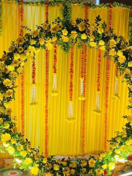 A beautiful circular backdrop for a Haldi ceremony, made with a wreath of yellow flowers and hanging floral tassels. The yellow fabric background enhances the bright and sunny theme.