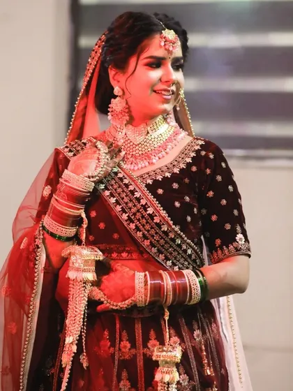 This portrait captures the bride in her beautiful maroon velvet lehenga, holding her kalire. The soft red lighting gives the image a warm, celebratory glow, highlighting her happy expression and the intricate details of her bridal attire.