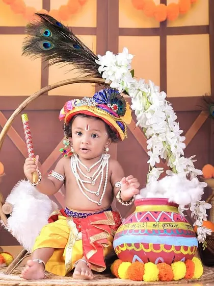 A baby dressed as Krishna, holding a flute and sitting next to a decorated pot, with a colorful headdress and peacock feathers.