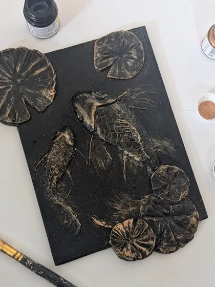 A top-down view of a koi fish textured art piece in progress. I love showing the process, with the paints and tools right next to the art. This piece uses layers of modeling paste to create the illusion of fish swimming beneath the water's surface.