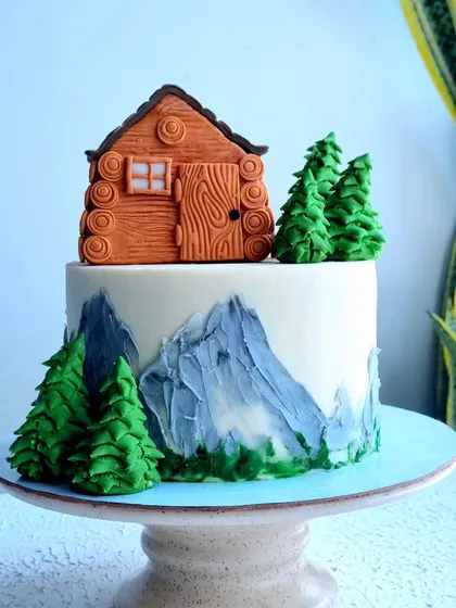A charming cottage-themed cake. This design features a log cabin topper, fondant pine trees, and buttercream mountains, creating a cozy, rustic scene.