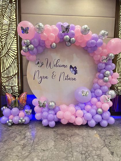 A beautiful butterfly-themed welcome backdrop for a party. The circular sign is personalized with the guests of honor's names and framed with a pink, purple, and silver balloon arch.