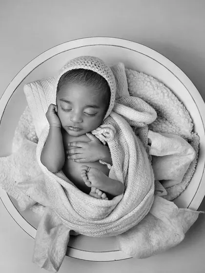 This black and white portrait shows a newborn curled up in a round bowl, wrapped in soft blankets. Monochrome photos emphasize the beautiful shapes and textures of the moment.