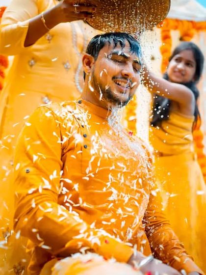 A joyful moment from a Haldi ceremony, capturing the groom being showered with rice and flowers under the bright sun.
