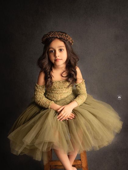 A simple, elegant portrait of a girl in an olive green dress and a golden headband, looking directly at the camera with a calm expression.